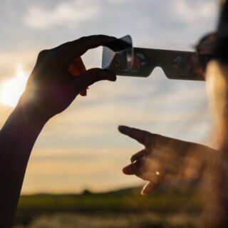 Person holds 3D glasses up to the sunset, lenses reflecting the sky as another hand points forward.
