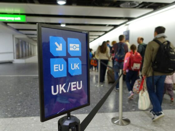 Airport sign with UK/EU icons directing travelers, foregrounded while a queue of passengers waits in the background.