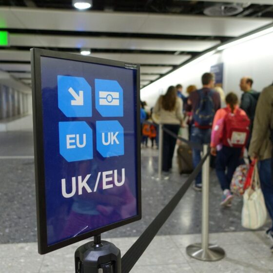 Airport sign with UK/EU icons directing travelers, foregrounded while a queue of passengers waits in the background.