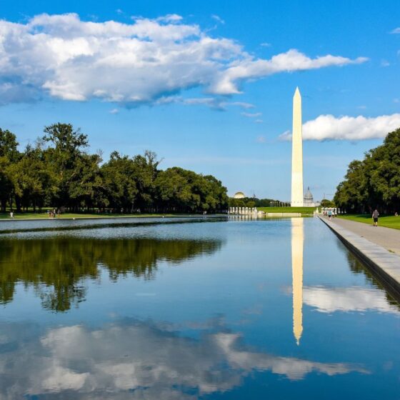 Washington Monument reflected in the Reflecting Pool with trees on both sides and a bright blue sky overhead.