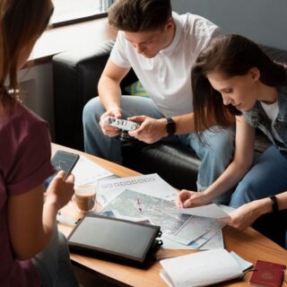 Three friends sit around a coffee table planning a trip, with maps, a passport, and devices visible.
