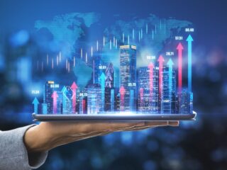 Hand holding a tablet projecting a holographic city skyline with blue and pink arrows and stock data above it (business/finance concept).