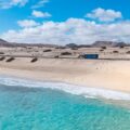 Bright blue sky over a sandy coastline with turquoise sea, a road, and a blue bus traveling along the dunes along the beach.