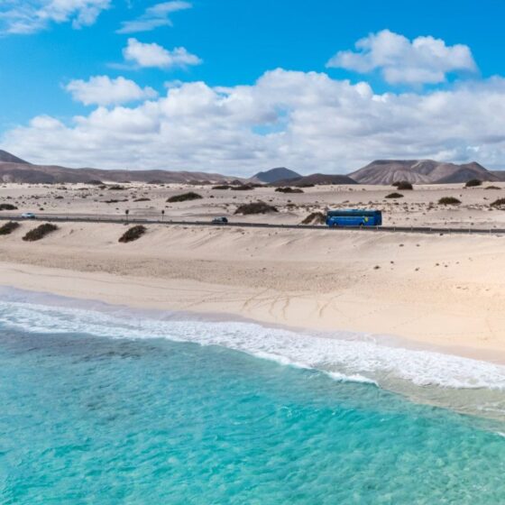Bright blue sky over a sandy coastline with turquoise sea, a road, and a blue bus traveling along the dunes along the beach.