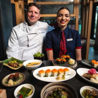 Two restaurant staff, a chef in a white coat and a server in a navy uniform, sit behind a table full of sushi dishes.",