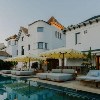 Upscale hotel courtyard with a rectangular pool, yellow umbrellas, and white loungers beside white buildings with red-tiled roofs.