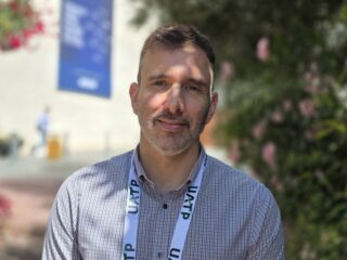 Portrait of a man with short hair and stubble wearing a checkered shirt and a UTAP lanyard, outdoors.