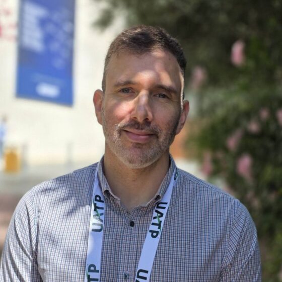 Portrait of a man with short hair and stubble wearing a checkered shirt and a UTAP lanyard, outdoors.