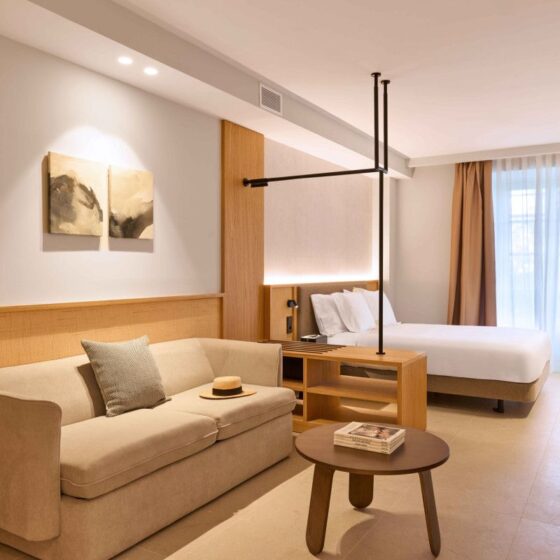 Hotel room with a beige sofa, round coffee table, and a king bed near a large window with brown drapes in a neutral, airy space.