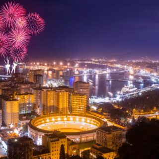 Night aerial view of a coastal city with pink fireworks over a brightly lit stadium and harbor lights.