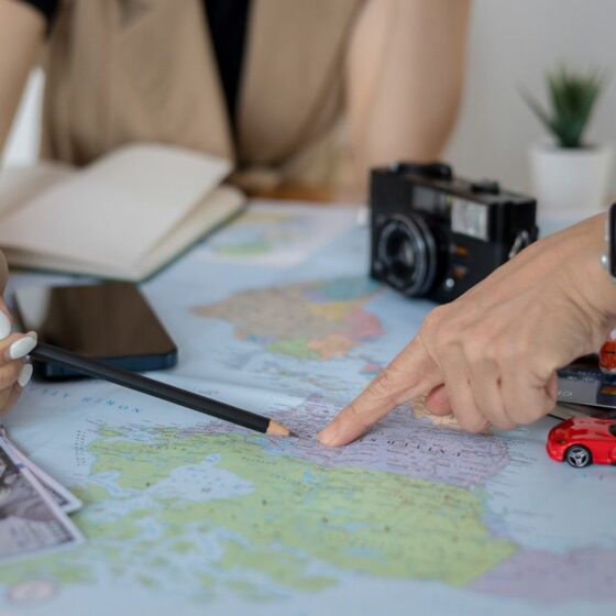 Two people plan a trip over a world map with a pencil, money, and travel gear nearby, signaling travel planning or budgeting.