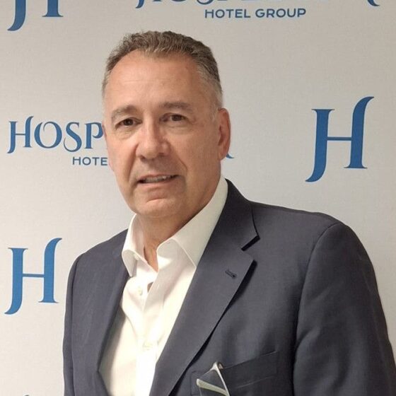 Portrait of a middle-aged man in a navy suit and white shirt, standing before a backdrop with blue hotel logos.
