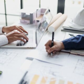 Two professionals collaborate at a desk with a laptop, plans, and a white safety helmet nearby