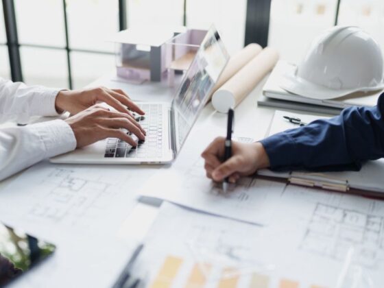 Two professionals collaborate at a desk with a laptop, plans, and a white safety helmet nearby