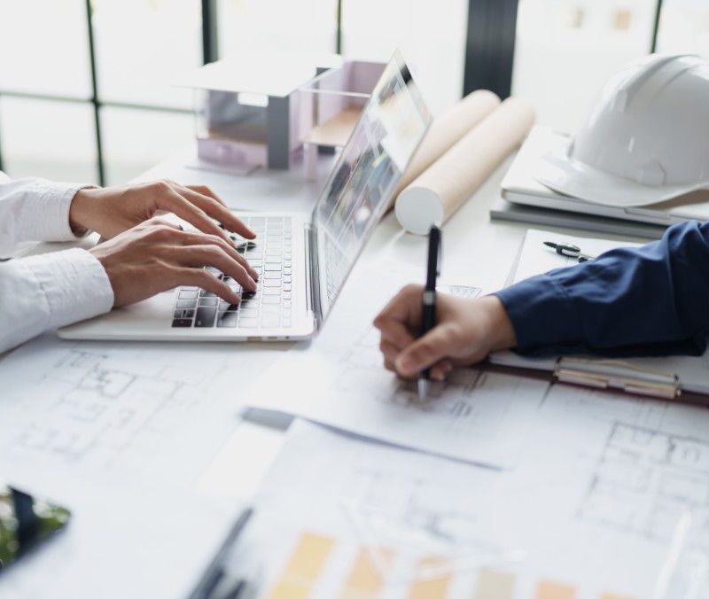 Two professionals collaborate at a desk with a laptop, plans, and a white safety helmet nearby