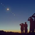 Silhouetted group of people on a hill watching a solar eclipse arc across a twilight sky.
