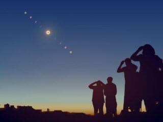Silhouetted group of people on a hill watching a solar eclipse arc across a twilight sky.