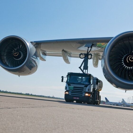 Tow tractor guiding a large commercial airplane under its wing on a sunny airport tarmac, with an orange cone in the foreground.