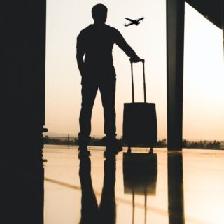 Silhouette of a traveler with a rolling suitcase at an airport doorway during sunset, plane visible in the distance.
