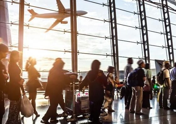 Passengers with rolling suitcases line up at an airport terminal, with a plane visible outside the glass wall as the sun glows.