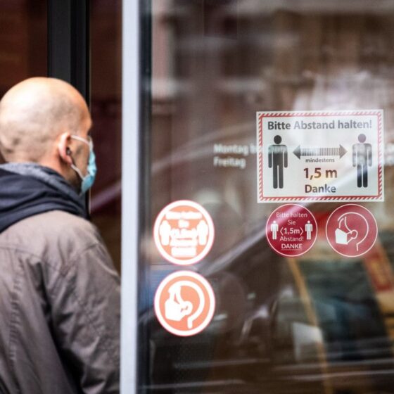 Person wearing a face mask standing beside a glass door with COVID-19 safety signs in German (Please keep distance).
