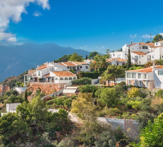 White hillside village with white-washed houses and orange-tiled roofs, greenery all around, under a clear blue sky and distant mountains in view.