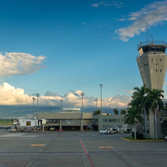 Airport tarmac with a tall control tower and terminal buildings under a blue sky with clouds.