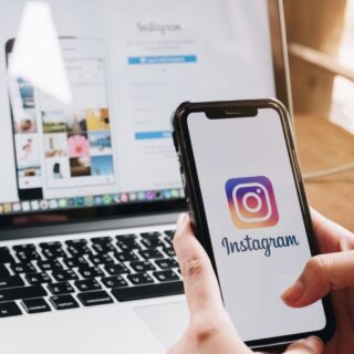 Person holds a smartphone showing the Instagram app logo, with a laptop displaying a photo grid in the background.