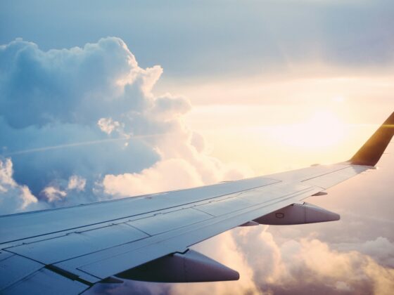 Airplane wing visible from the window with blue sky, fluffy clouds, and warm sunset light.