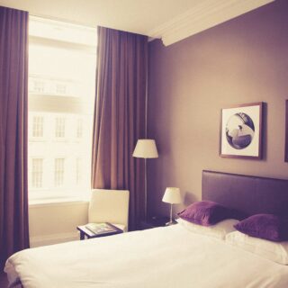 Hotel bedroom with two beds, white linens, purple pillows, and wall art; large window with brown curtains on the left.