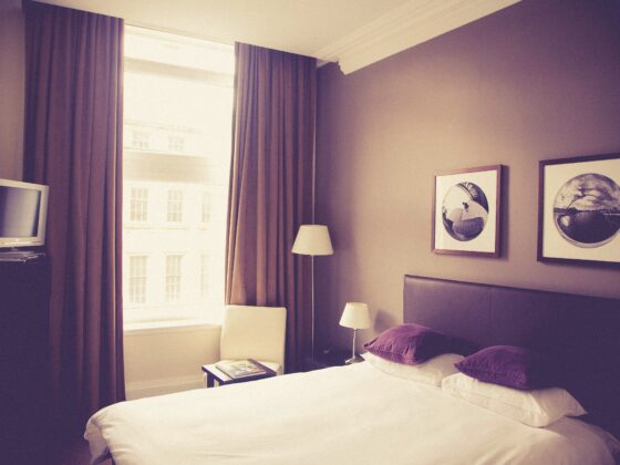 Hotel bedroom with two beds, white linens, purple pillows, and wall art; large window with brown curtains on the left.