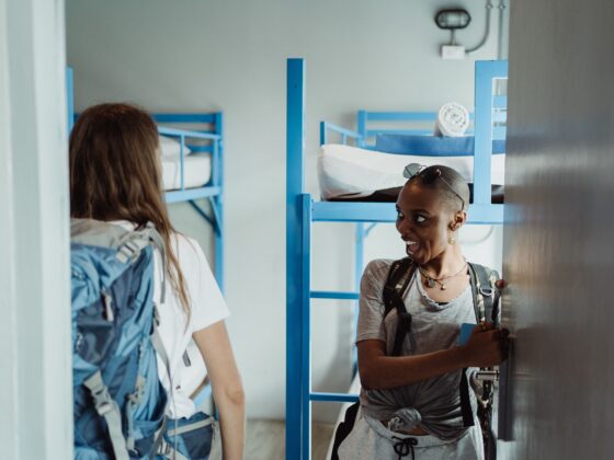 Two friends standing in a dorm doorway with blue metal bunk beds in the background; one wearing sunglasses on her head and a backpack smiles at the other.