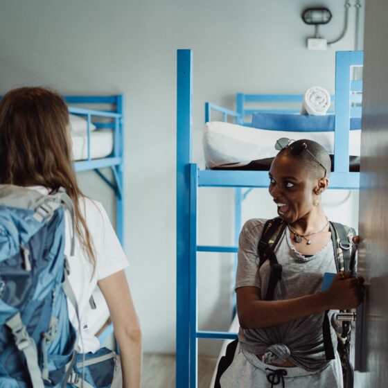 Two friends standing in a dorm doorway with blue metal bunk beds in the background; one wearing sunglasses on her head and a backpack smiles at the other.