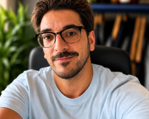 Portrait of a man with dark hair and glasses, wearing a light blue T-shirt, seated at a desk with bookshelves in the background.