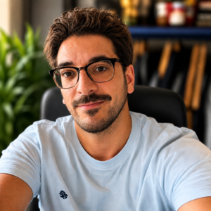 Portrait of a man with dark hair and glasses, wearing a light blue T-shirt, seated at a desk with bookshelves in the background.