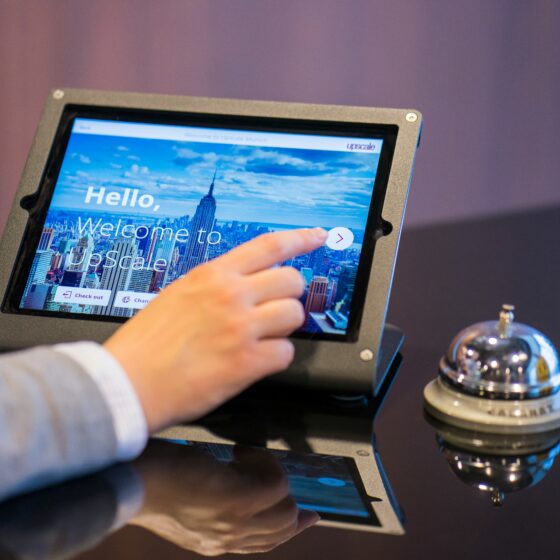 Person taps a tablet-like kiosk showing a city skyline and the words 'Hello, Welcome to Upscale' on the screen.