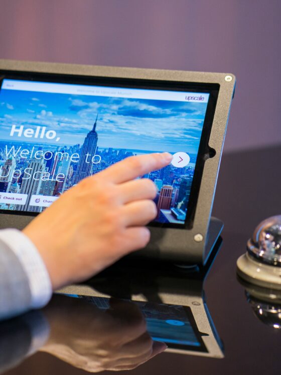 Person taps a tablet-like kiosk showing a city skyline and the words 'Hello, Welcome to Upscale' on the screen.